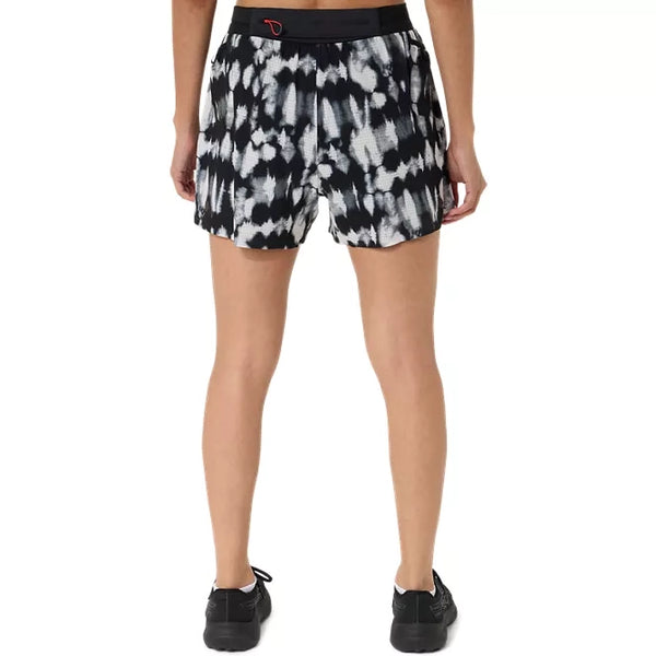 En person står med ryggen til, iført sorte sneakers og ASICS Limited Series Metarun Split Shorts til kvinder i farvekombinationen Cream/Performance Black – et stilfuldt par løbeshorts til kvinder med et sort-hvidt tie-dye-mønster og en kontrastfarvet sort linning. Hænderne hviler ned langs siden på en ensfarvet hvid baggrund.