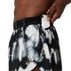 En person lægger en lille hvid genstand, muligvis et kort eller et stykke papir, ned i lommen på et par ASICS Limited Series Metarun Split Shorts til mænd i farven Cream/Performance Black, mens vedkommendes overkrop er delvist synlig.