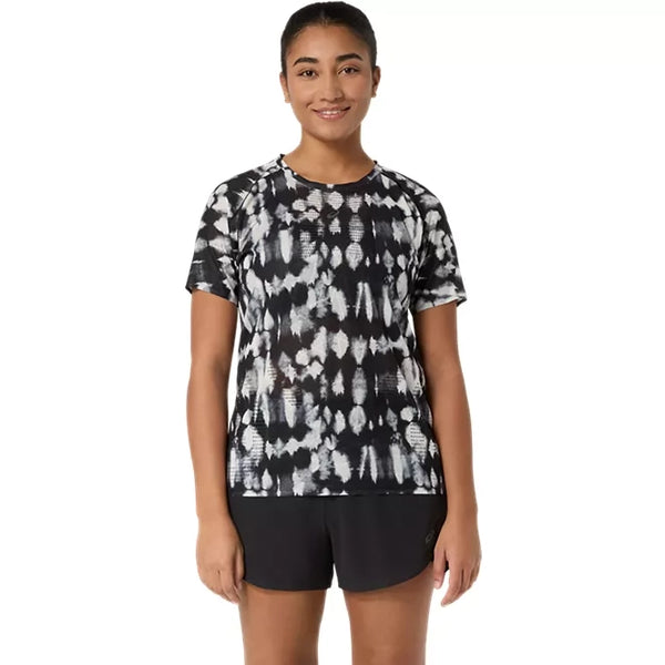 En kvinde står med ansigtet vendt fremad og smiler. Hun har en Asics Limited Series Metarun SS Top til kvinder i farven Cream/Performance Black på – en løbetrøje til kvinder med den innovative ACTIBREEZE-teknologi, der sikrer bedre åndbarhed – kombineret med ensfarvede sorte shorts. Baggrunden er helt hvid, og hendes arme hænger afslappet ned langs siden.