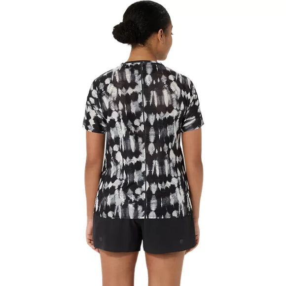 En person står med ryggen til og har en Asics Limited Series Metarun Ss Top til kvinder i farven Cream/Performance Black samt sorte shorts på. Håret er sat op i en knold, og baggrunden er enkel og overskuelig.