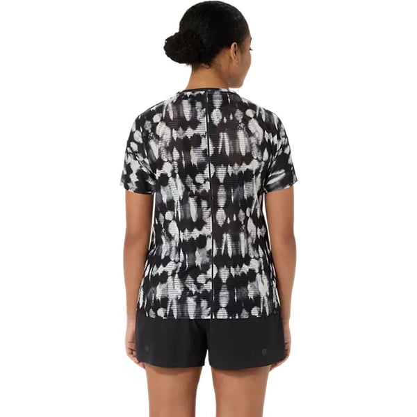 En person står med ryggen til og har en Asics Limited Series Metarun Ss Top til kvinder i farven Cream/Performance Black samt sorte shorts på. Håret er sat op i en knold, og baggrunden er enkel og overskuelig.