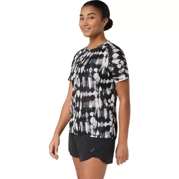 En kvinde står iført en ASICS Limited Series Metarun SS Top, Dame, i farven Cream/Performance Black – en stilfuld løbetrøje til kvinder med den innovative ACTIBREEZE-teknologi – kombineret med sorte sportsshorts. Foran en ensfarvet hvid baggrund står hun med blikket let vendt til siden og smiler blidt.