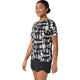 En kvinde står iført en ASICS Limited Series Metarun SS Top, Dame, i farven Cream/Performance Black – en stilfuld løbetrøje til kvinder med den innovative ACTIBREEZE-teknologi – kombineret med sorte sportsshorts. Foran en ensfarvet hvid baggrund står hun med blikket let vendt til siden og smiler blidt.