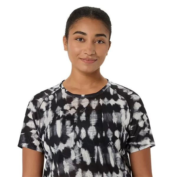 En person med mørkt hår, der er sat op, iført en Asics Limited Series Metarun SS Top til kvinder i farven Cream/Performance Black – en løbetrøje til kvinder med avanceret ACTIBREEZE-teknologi – står og smiler let foran en ensfarvet hvid baggrund.