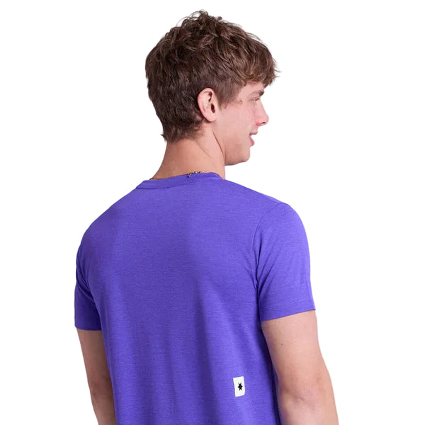 En person med kort brunt hår ser væk fra kameraet og er iført en almindelig lilla Saysky Logo Combat Pro T-shirt, Herre. Et lille hvidt mærke med et sort symbol ses nederst på ryggen. Baggrunden er hvid.