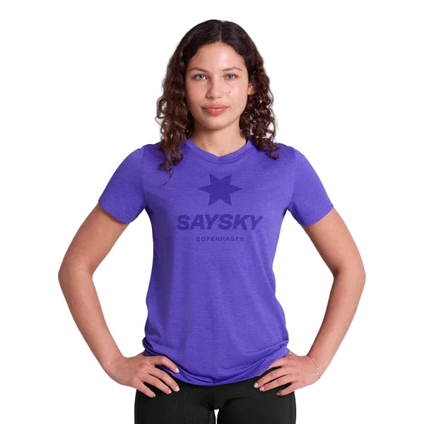 En kvinde med krøllet brunt hår står med hænderne på hofterne, iført en lilla Saysky Logo Combat Pro T-shirt, Dame og sorte bukser på en almindelig hvid baggrund.