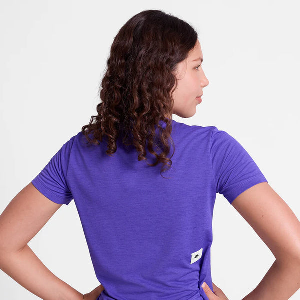 En kvinde med krøllet brunt hår ses bagfra iført en kortærmet lilla Saysky Logo Combat Pro T-shirt (Dame). Et lille rektangulært hvidt mærke er synligt nederst på ryggen. Baggrunden er helt hvid.