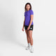 En person står på en almindelig hvid baggrund iført en Saysky Logo Combat Pro T-shirt, Dame, Purple, sammen med sorte shorts, hvide sokker og hvide sportssko. Vedkommende kigger fremad og smiler let.