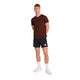 En person står foran en hvid baggrund iført en rød Saysky Logo Pace T-shirt (herre), sorte shorts med et mærke, hvide sokker og hvide sneakers. Armene er placeret bag ryggen.