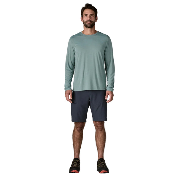 En mand står og kigger fremad iført en blå, salviefarvet Patagonia Long-Sleeved Capilene Cool Shirt, mørkeblå shorts og sorte sportssko med orange såler på en almindelig hvid baggrund.
