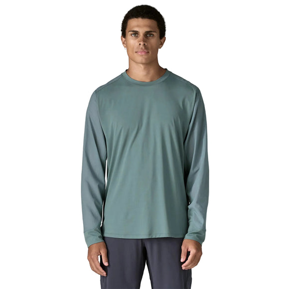 En mand med kort, krøllet hår og et neutralt udtryk står og kigger fremad i en Patagonia Long-Sleeved Dirt Craft Jersey, Herre, Blue Sage, sammen med mørke bukser mod en almindelig hvid baggrund og viser trailrunning-stil.