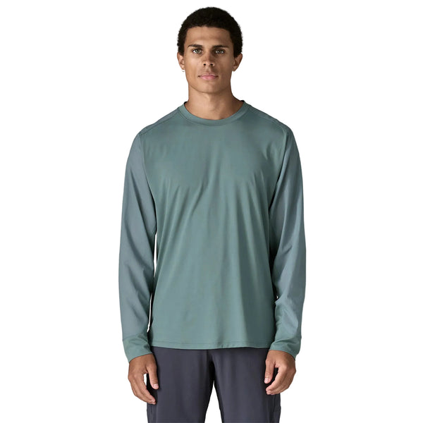 En mand med kort, krøllet hår og et neutralt udtryk står og kigger fremad i en Patagonia Long-Sleeved Dirt Craft Jersey, Herre, Blue Sage, sammen med mørke bukser mod en almindelig hvid baggrund og viser trailrunning-stil.