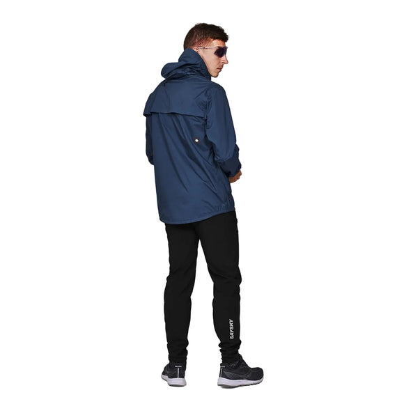 En person iført Saysky Element 3L Waterproof Jacket, Herre, i blå står med ansigtet vendt væk fra kameraet mod en almindelig lysegrå baggrund. Vedkommende har også sorte bukser, mørke sko og solbriller på.