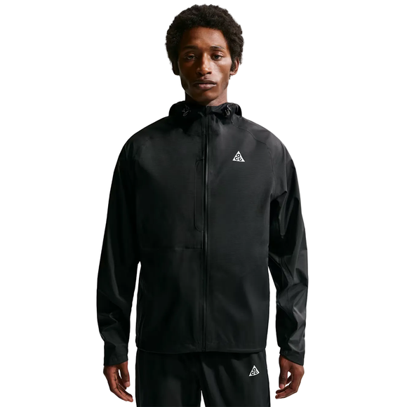 En mand ser frem for sig iført en sort Nike ACG Sfadv Cosmic Peaks Jkt, Herre, Black/Summit White, parret med matchende sorte bukser med små trekantede logoer på en almindelig lys baggrund.