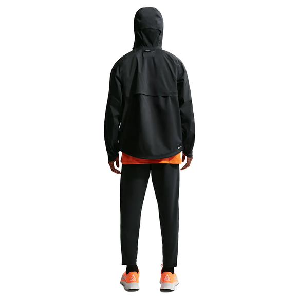 En person kigger væk iført Nike ACG Sfadv Cosmic Peaks Jkt, Herre, Black/Summit White, sammen med sorte bukser, en orange skjorte indenunder og hvide sneakers med orange detaljer mod en almindelig lys baggrund.