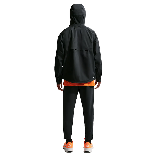En person kigger væk iført Nike ACG Sfadv Cosmic Peaks Jkt, Herre, Black/Summit White, sammen med sorte bukser, en orange skjorte indenunder og hvide sneakers med orange detaljer mod en almindelig lys baggrund.