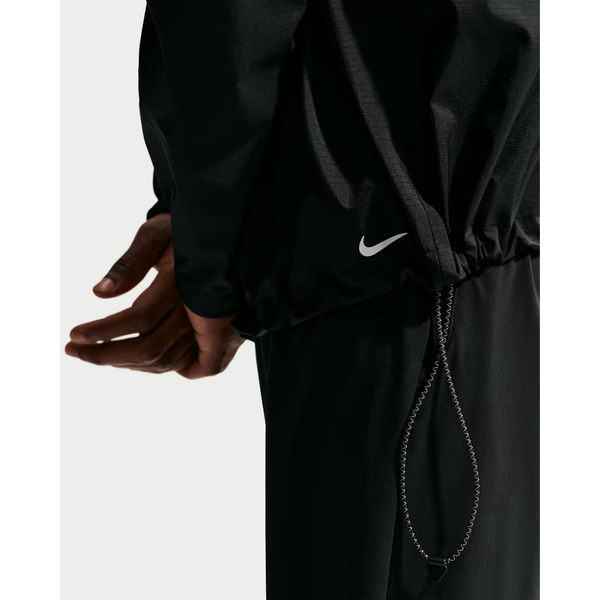 Nærbillede af en person iført Nike ACG Sfadv Cosmic Peaks Jkt til mænd i Black/Summit White, med justerbar snøre forneden og en del af hånden og armen på en almindelig baggrund.