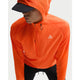 En person i en lys Safety Orange Nike ACG Trail Aireez Jkt med lynlås og lille logo på brystet samt en sort kasket kigger nedad. Den vandafvisende letvægtsjakke skiller sig ud fra den almindelige lyse baggrund.