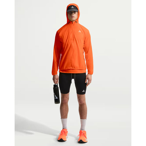 En person står med ansigtet fremad i Nike ACG Trail Aireez Jkt (Safety Orange/Summit White), sorte shorts, hvide sokker, orange løbesko, sort kasket og med en sort vandflaske i hånden. Den vandafvisende jakke skiller sig ud fra den ensfarvede, lyse baggrund.