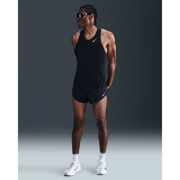En person iført Nike M Nk Dfadv Aeroswift Jakob Singlet, Herre, Black/Metallic Gold med sorte løbeshorts, hvide sneakers og solbriller står med armene bag ryggen mod en mørk baggrund.