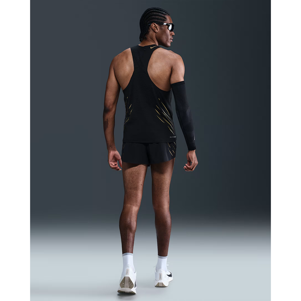 En mand i Nike M Nk Dfadv Aeroswift Jakob Singlet til mænd (Black/Metallic Gold), sorte shorts, hvide sokker og løbesko står med ansigtet vendt væk. Han har flettet hår, solbriller, et sort armlæn, og baggrunden er almindelig mørk.