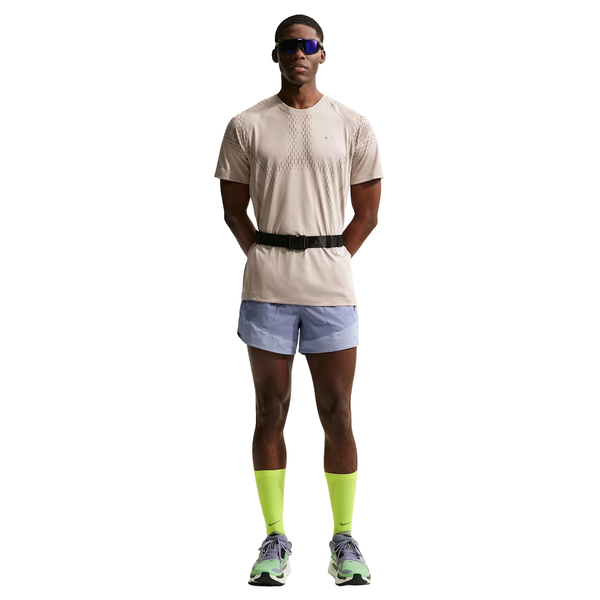 En person, der ser fremad med solbriller, er iført en Nike Stride Novelty SS Løbetop (creme/minkbrun/reflekterende sølv), lyseblå løbeshorts, sort taljebælte, gule knæstrømper og løbesko med grøn og lilla accent.