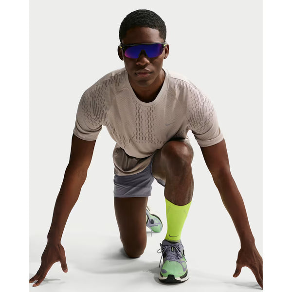 En person er iført Nike Stride Novelty SS Running Top, Herre, Cream Ii/Mink Brown/Reflective Silv, grå shorts, neongule sokker, løbesko og reflekterende solbriller i en sprinters startposition på en hvid baggrund.