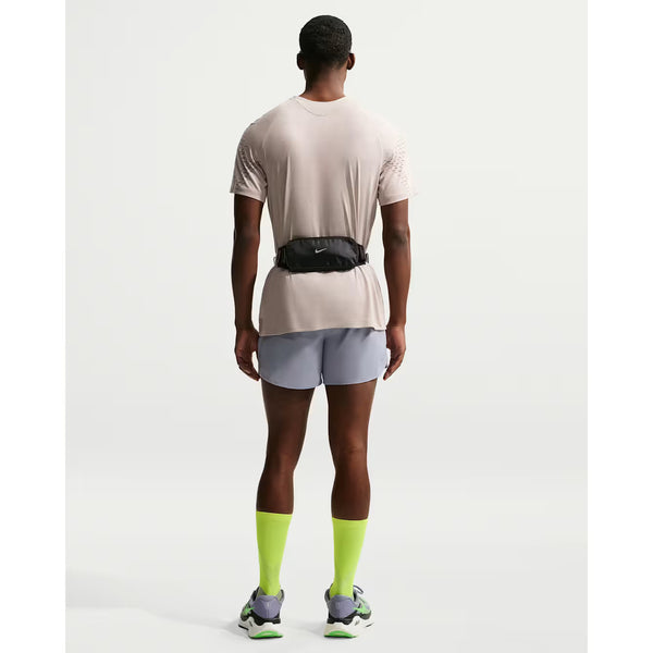 En person kigger væk fra kameraet iført Nike Stride Novelty SS Running Top i Cream Ii/Mink Brown/Reflective Silv, lysegrå shorts, neongule-grønne sokker, hvide løbesko og en lille sort bæltetaske. Baggrunden er helt hvid.