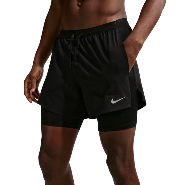 En person er iført sorte Nike Stride 5" Hybrid Running Shorts med indbygget fugttransporterende kompressionsfor, med den ene hånd i en lomme. Nike-logoet ses på venstre ben. Kun underkroppen og overdelen af benene er synlige.
