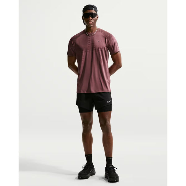 En person står med front mod kameraet i en rødbrun sportstrøje, sorte sokker, sorte sko, solbriller, en sort baglæns kasket og Nike Stride 5" Hybrid Running Shorts til mænd (Black/Reflective Silver) på en almindelig hvid baggrund.