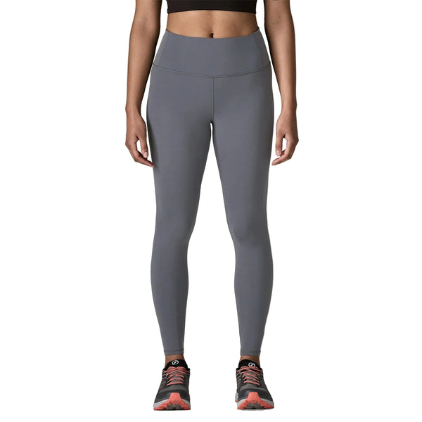 En person iført Patagonia Mas Maipo Tights, Dame, i Smolder Blue, en sort crop top og sorte og pink sportssko står på en hvid baggrund med afslappede arme langs siden.