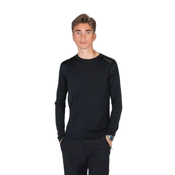 En ung mand med kort brunt hår bærer Fusion Merino 150 LS langærmet sort skjorte og sorte bukser, mens han står med hænderne i lommerne på en almindelig hvid baggrund.