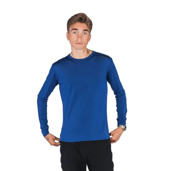 En ung mand med kort brunt hår står på en almindelig hvid baggrund iført Fusion's Merino 150 LS, Herre, Night Blue top og sorte bukser. Hans hænder hviler på hofterne, og han har et neutralt ansigtsudtryk.