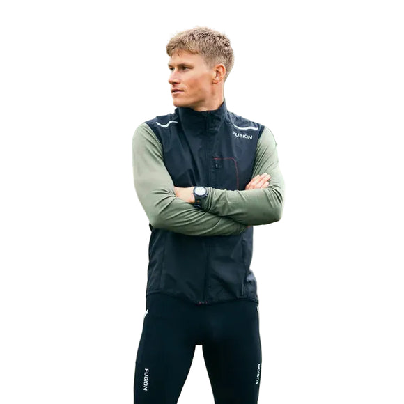 En person med kort blondt hår står udendørs på græs iført Fusion S1 Run Vest, Herre, Black over en grøn langærmet skjorte, sorte leggings og et ur. Slørede træer er synlige i baggrunden. Armene er krydsede.