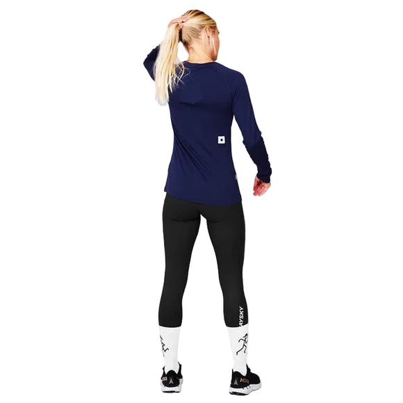 En kvinde med langt blondt hår ses bagfra indendørs iført Saysky W Merino Base 165 Long Sleeve, Dame, Blue shirt med sorte leggings, hvide sokker og sorte sportssko.