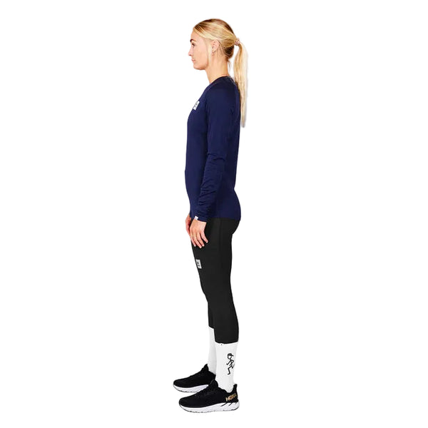 En blond kvinde med hestehale står i profil mod en grå baggrund iført Saysky W Merino Base 165 Long Sleeve, Dame, Blue shirt, sorte leggings, hvide sokker og sorte sportssko.