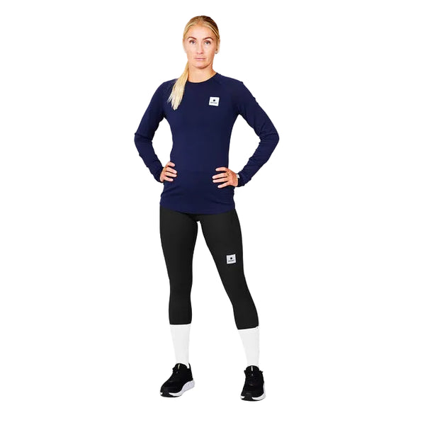 En kvinde står med ansigtet fremad og hænderne på hofterne, iført Saysky W Merino Base 165 Long Sleeve, Dame, Blue, sammen med sorte og hvide leggings og sorte sneakers på en almindelig neutral baggrund.