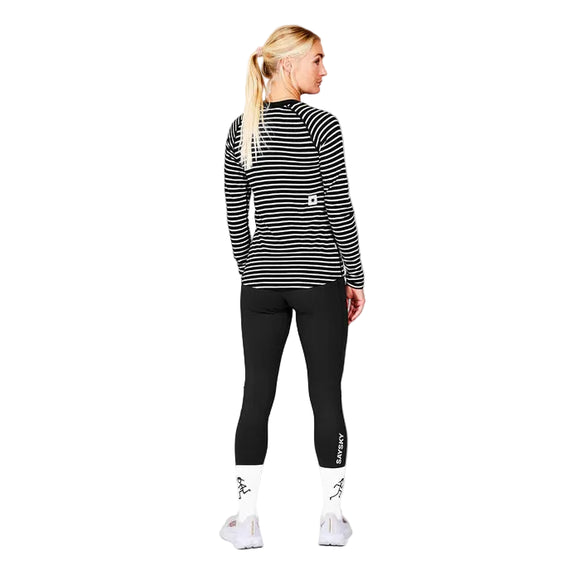 En kvinde med blondt hår i hestehale kigger væk, iført Saysky W Merino Base 165 Long Sleeve, Dame, Stripes top med sorte leggings, hvide sokker og hvide sneakers på en almindelig grå baggrund.