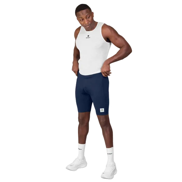 En mand står foran en ensfarvet hvid baggrund iført Saysky Mesh Base Layer Singlet, Herre, i hvid. Han har den på uden på et andet åndbart mesh-undertøj, kombineret med marineblå cykelshorts, hvide sokker og hvide sportssko. Han kigger nedad og retter på sine shorts med begge hænder og er dermed et levende eksempel på den funktionelle stil inden for moderne herresportstøj.