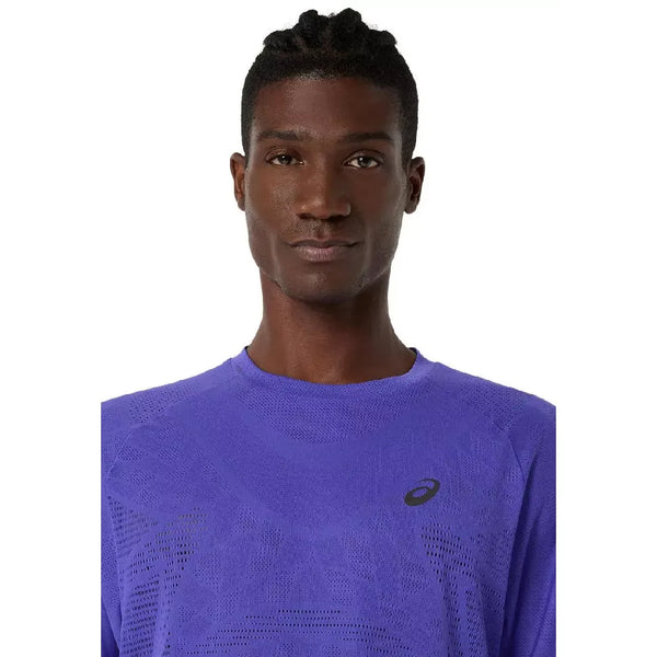 En person med kort, flettet hår er iført en koboltblå Asics Metarun Ss Top, Herre, med et sort Asics-logo på venstre bryst, og ser direkte ind i kameraet mod en almindelig hvid baggrund.