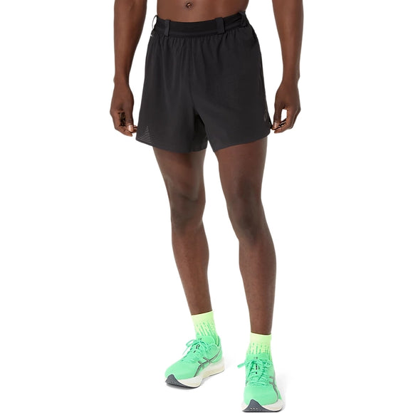 En person står med ansigtet vendt fremad iført et par Asics Metarun 5In Shorts, Herre, Performance Black, kombineret med en matchende sort top, neongrønne sokker og lysegrønne løbesko. Baggrunden er hvid. Personens ansigt kan ikke ses på billedet. Shortsene byder på den elegante ydeevne, der kendetegner Asics’ premium-løbeshorts til herrer.
