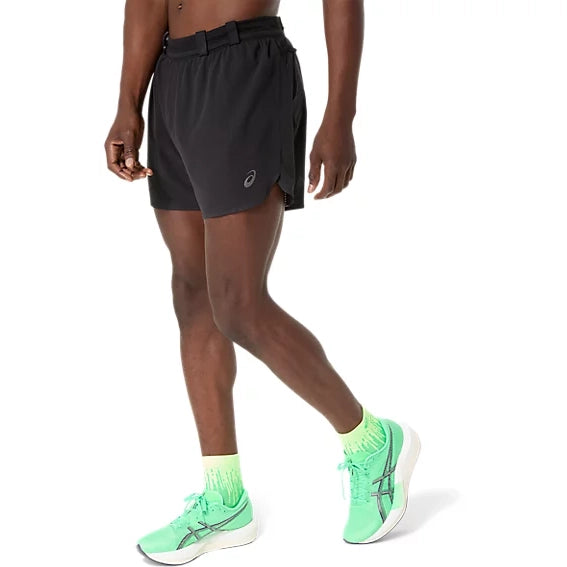 En person har Asics Metarun 5In Shorts, Herre, Performance Black på, som er udstyret med ACTIBREEZE-teknologi for forbedret åndbarhed, samt lysegrønne sokker og lysegrønne løbesko med sorte detaljer, og går fremad foran en ensfarvet hvid baggrund.