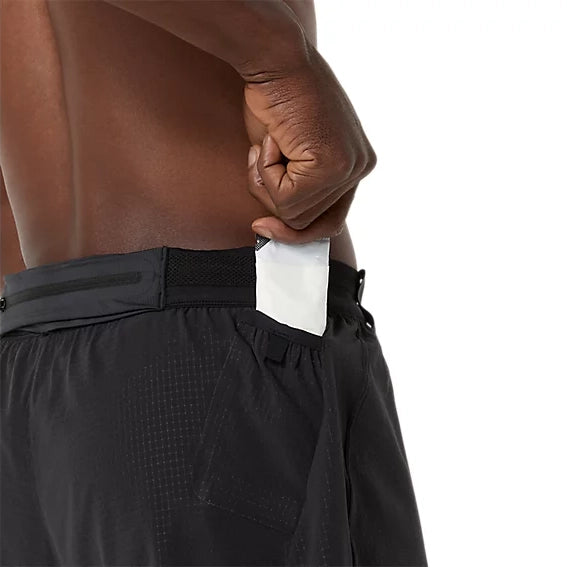 Man ser en person, der bærer ASICS Metarun 5In Shorts, Herre i farven Performance Black, sætte en hvid, rektangulær genstand – muligvis en telefon – ned i sidelommen med lynlås. Billedet viser et nærbillede af personens overkrop og hånd på en ensfarvet hvid baggrund.