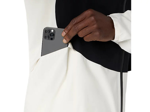 En person iført en Asics Metarun Legend-jakke, herre, creme/Performance Black – en populær vandtæt løbejakke – lægger en grå smartphone med tre bagkameraer ned i en sidelomme.