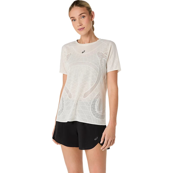 En kvinde står med ansigtet vendt fremad foran en ensfarvet hvid baggrund. Hun har en cremefarvet ASICS Metarun Ss-top til kvinder med mesh-detaljer og sorte shorts på. Den ene arm hænger afslappet ned langs siden, mens den anden er let bøjet.