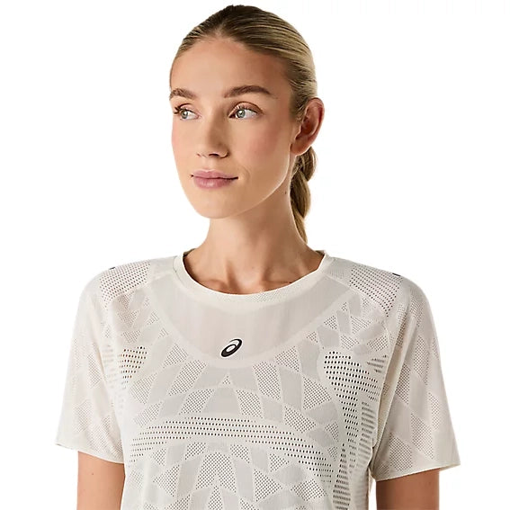En kvinde med blondt hår, der er sat op, har en Metarun SS Top, Dame, Cream fra Asics på – en hvid løbetrøje med et geometrisk mønster og det ikoniske Asics-logo. Hun kigger let mod venstre med et neutralt udtryk foran en ensfarvet hvid baggrund. Denne Metarun SS Top forener på smukkeste vis ydeevne og stil til den moderne løber.