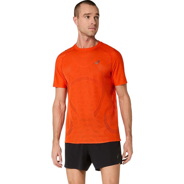 En person står foran en hvid baggrund iført en Metarun Ss Top til mænd i farven »cherry tomato« fra Asics og sorte shorts, mens vedkommende kigger lidt til siden.