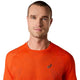 En mand med kort hår har en orange Asics Metarun Ss Top, Herre, Cherry Tomato på, der er prydet med det ikoniske Asics-logo. Tøjet er fremstillet med den innovative ACTIBREEZE-teknologi, og han står med ansigtet vendt fremad og et neutralt udtryk på en ensfarvet hvid baggrund.