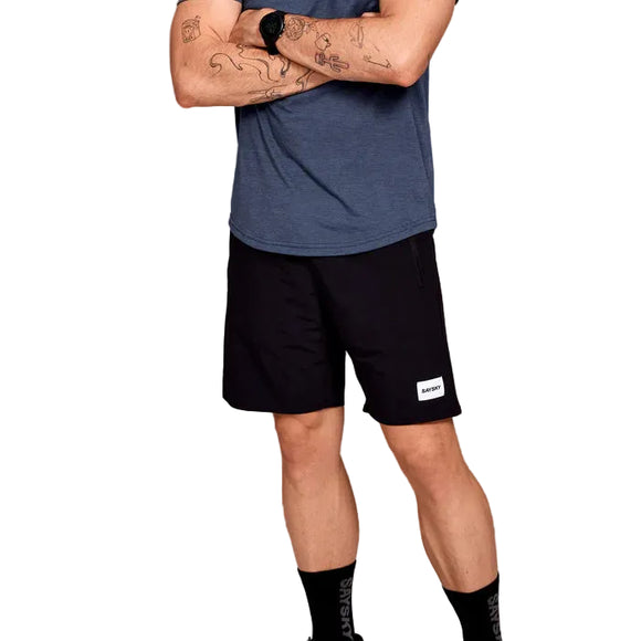 En herre står med korslagte arme, viser tatoveringer og er iført en blå t-shirt, sorte Saysky Motion Running Shorts 8", sorte sokker og et smartwatch. Løbeshortsene har et hvidt rektangulært mærke. Billedet er beskåret ved skuldrene.