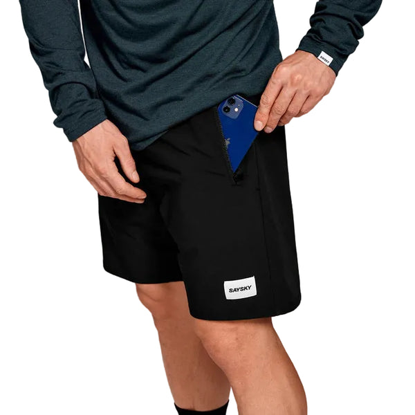 En person i en mørk langærmet skjorte og sorte Saysky Motion Running Shorts 8" (herre) putter en blå smartphone i shortsens lynlåslomme foran. Personens ansigt er ikke synligt.